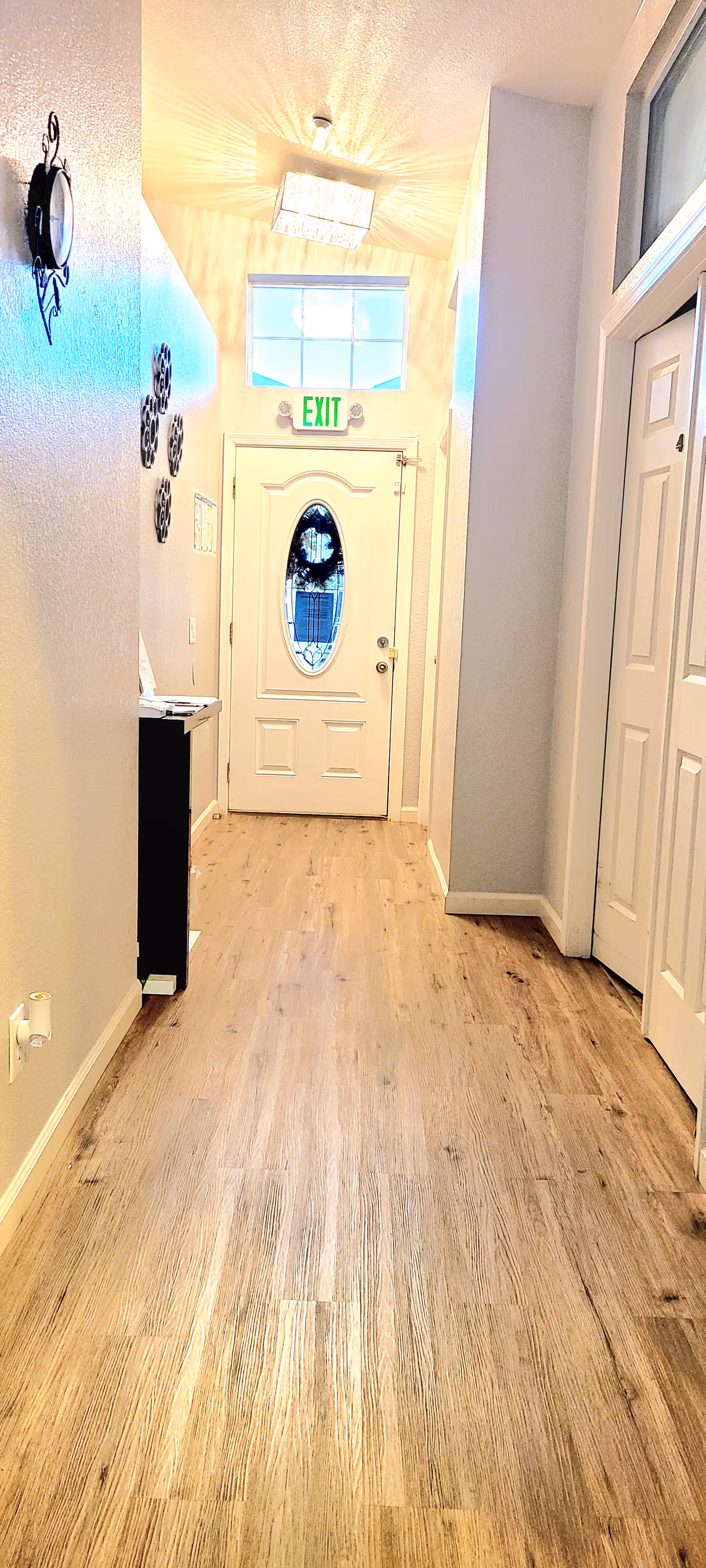 A bright hallway with wooden flooring leading to a white door with an oval glass window. Above the door is a green exit sign and a rectangular window letting in natural light. The walls are light-colored, and there is a decorative wall clock and wall art on the left side. On the right side, there are white closet doors and a window above them. A modern ceiling light fixture is visible near the door.