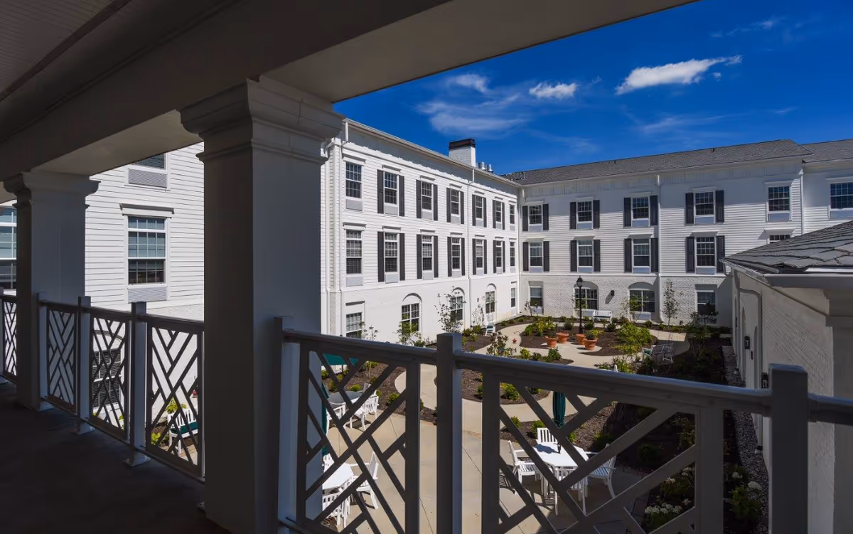 View of a bright, sunny courtyard garden area surrounded by a white multi-story building with many windows. The courtyard has paved walkways, potted plants, and white outdoor tables and chairs. The photo is taken from a covered balcony with white railings and columns.