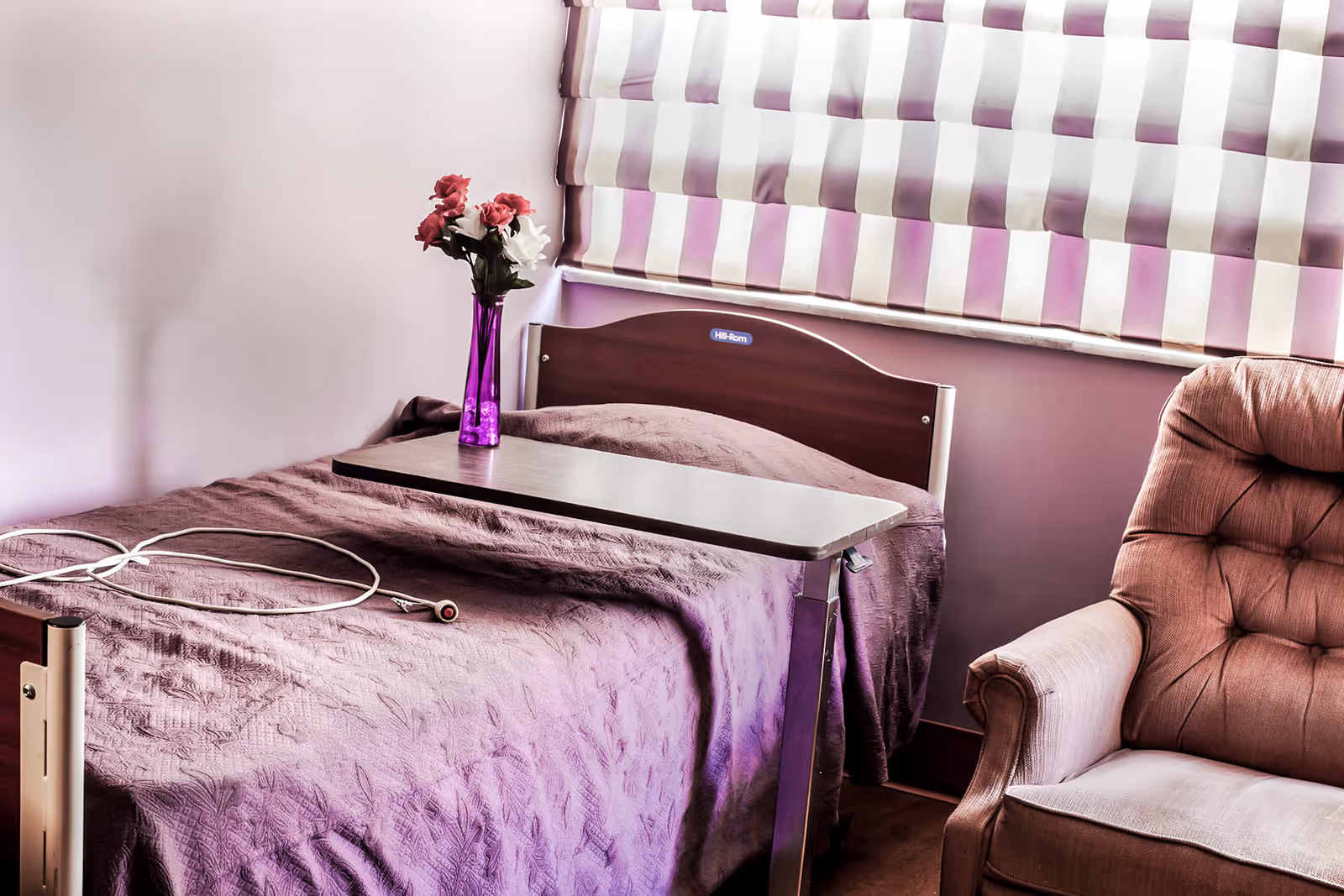 A senior living facility bedroom with a hospital-style bed covered in a purple blanket, a purple vase with pink and white flowers on an overbed table, a cushioned armchair, and a window with purple and white striped blinds.