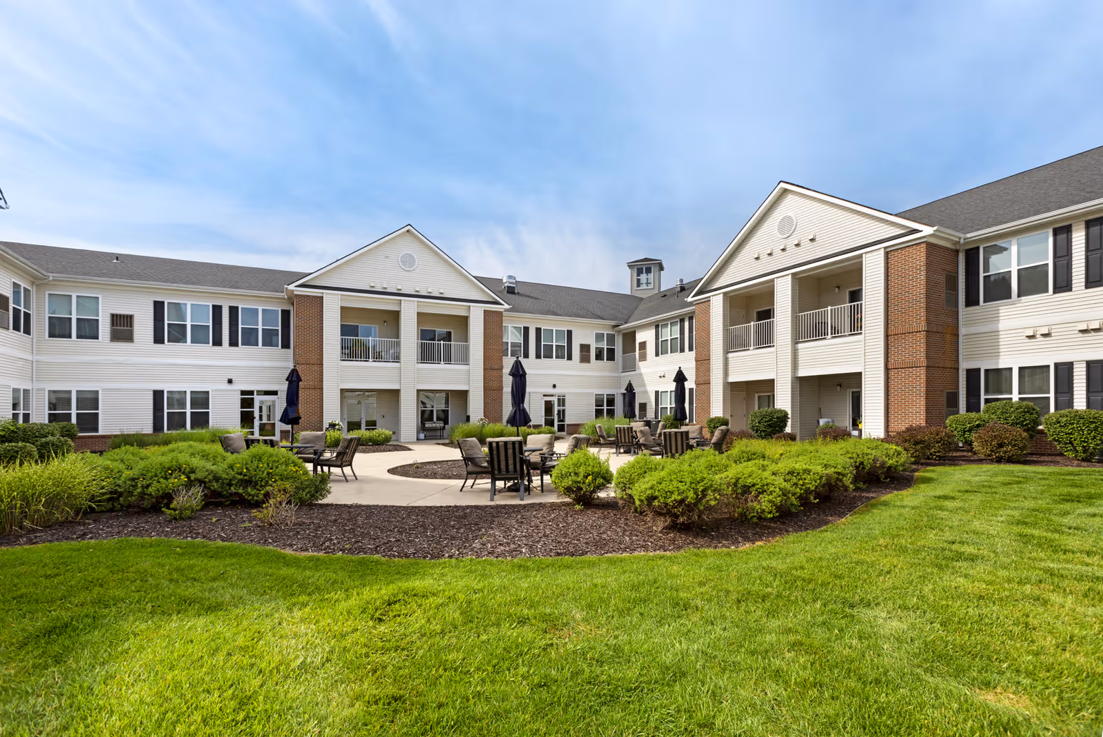 Outdoor courtyard area of a senior living facility with green grass, shrubs, and a circular paved seating area with chairs and umbrellas. The building surrounding the courtyard is two stories with white siding, brick accents, and balconies.