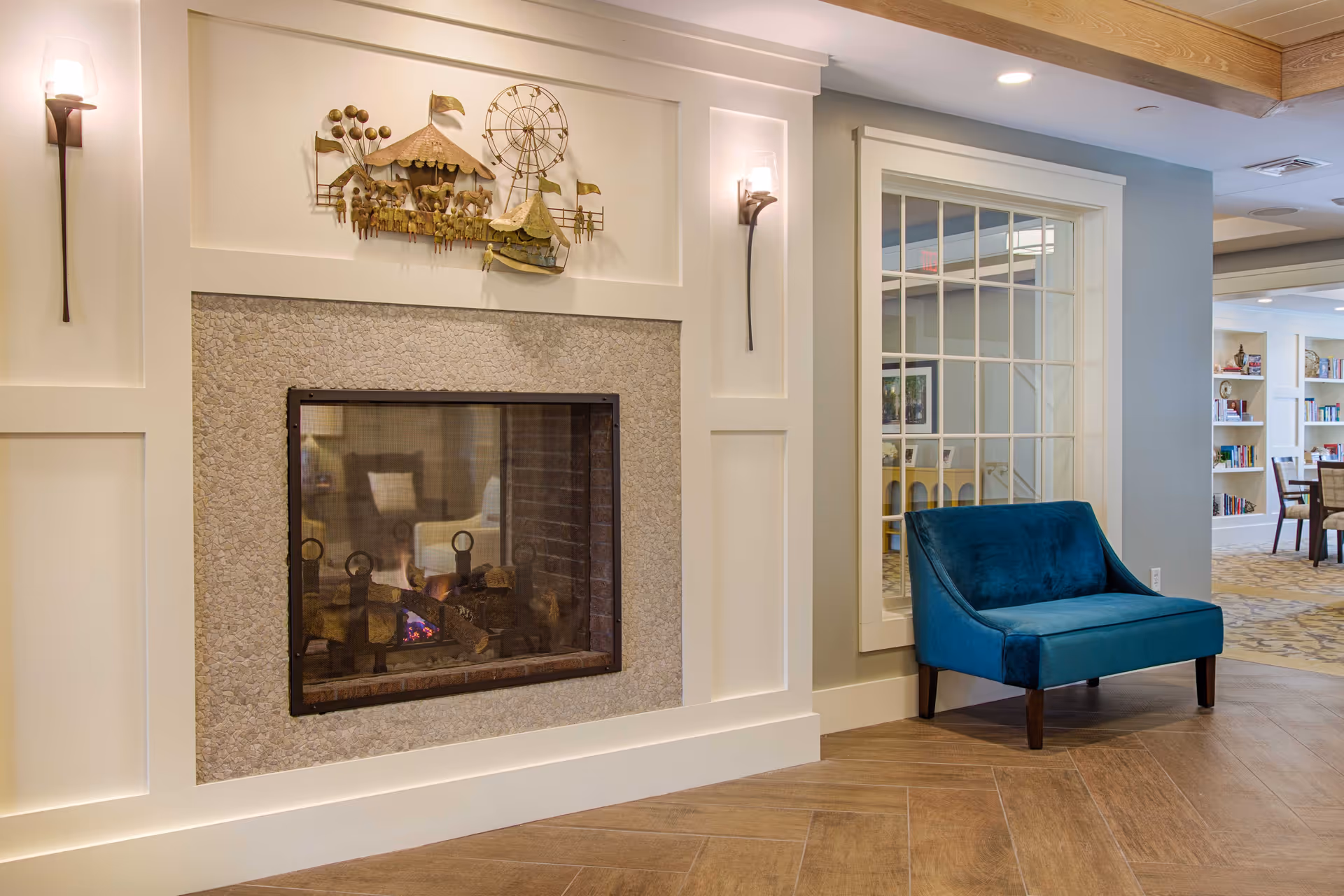 A cozy interior corner featuring a modern fireplace with a stone surround and a decorative metal wall art above it. On the right side, there is a blue upholstered chair placed against a wall with a large window that looks into another room with bookshelves and dining tables. The floor is wooden with a herringbone pattern, and the walls are painted in soft neutral tones with recessed lighting and wall sconces.