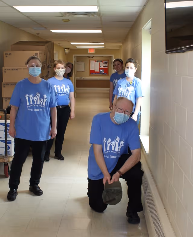 Five people wearing blue 'Hands Caring Together' t-shirts and face masks stand and kneel in a hallway with beige walls and a tiled floor. One person is kneeling near a window on the right side, holding a hat. There are stacked cardboard boxes on the left side of the hallway.