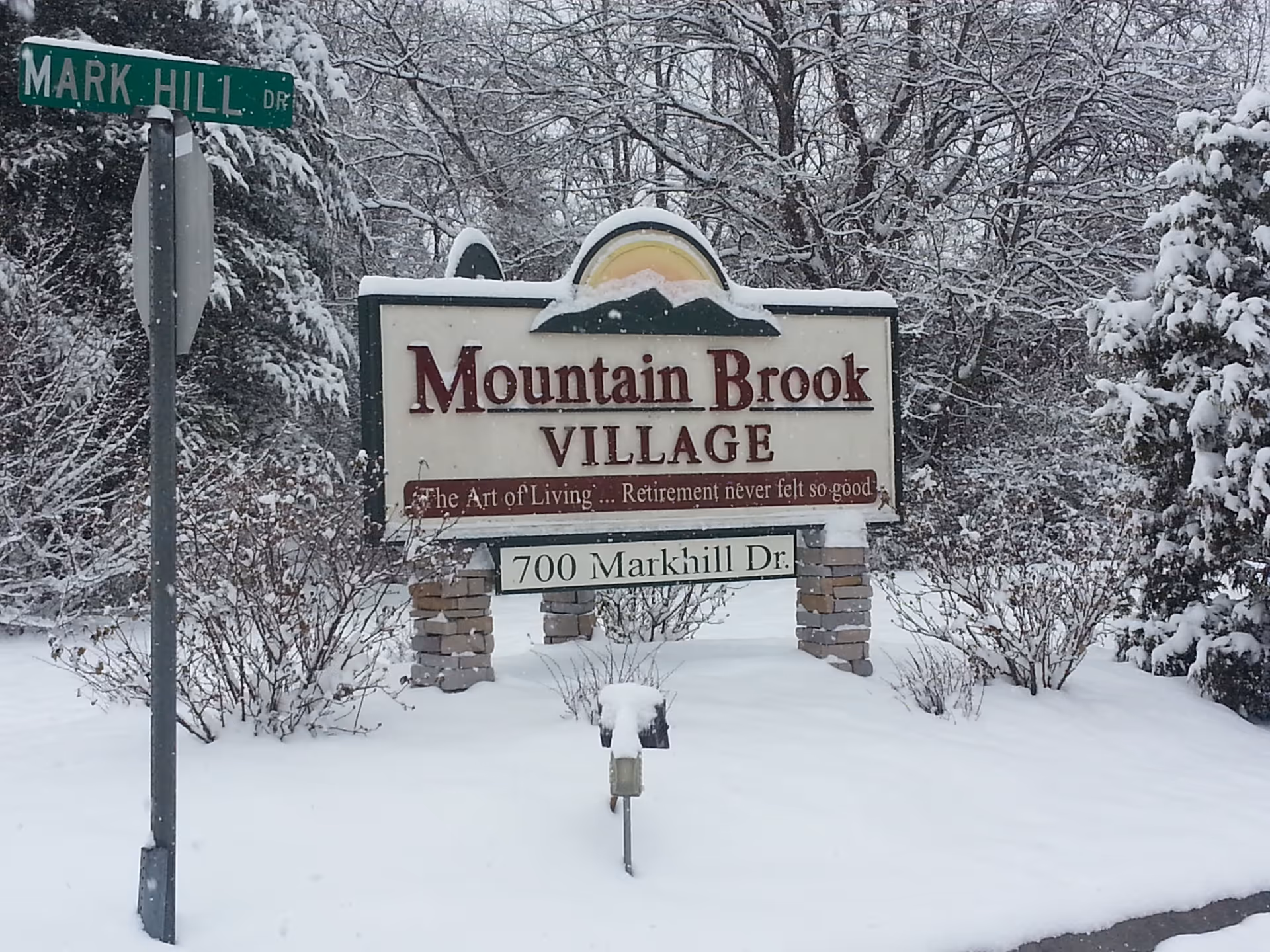 Snow-covered entrance sign that reads 'Mountain Brook Village' and '700 Markhill Dr' beside a Mark Hill Dr street sign.