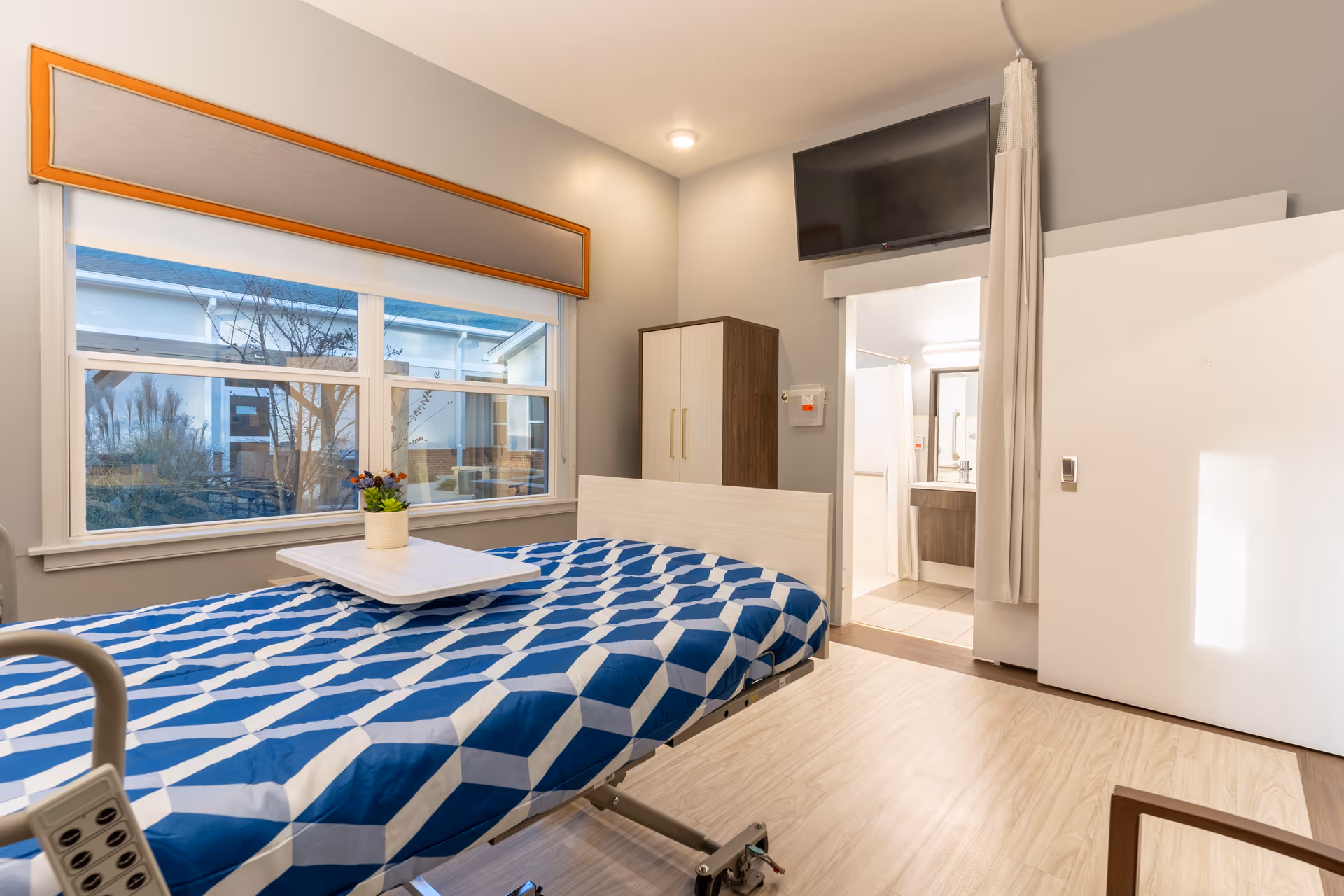 A bright and clean patient room in a rehab facility featuring a hospital bed with a blue and white geometric patterned blanket, a small table with a potted plant on the bed, a large window with an orange and gray valance, a wardrobe, a wall-mounted TV, and an open door leading to a bathroom.