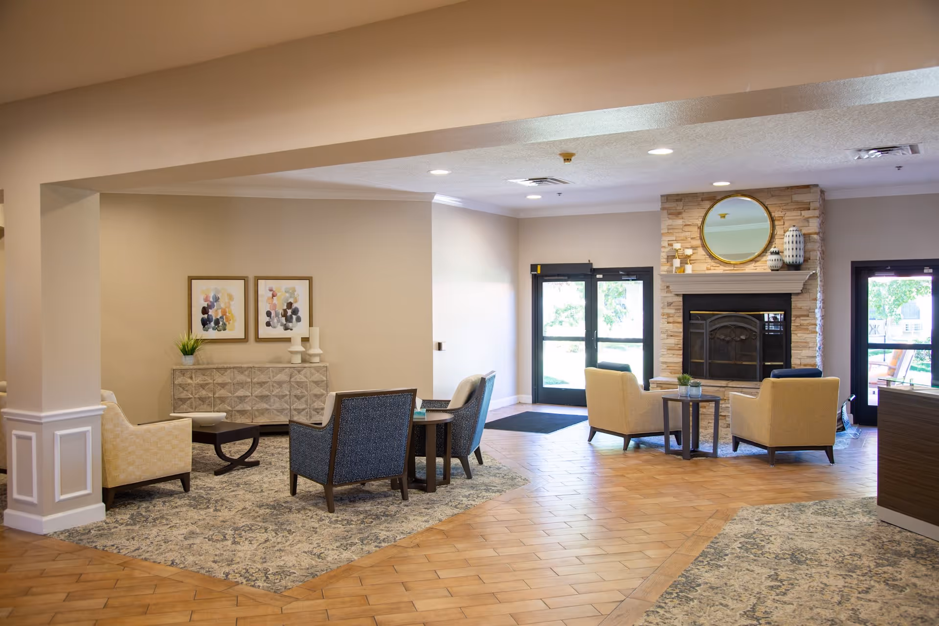 A spacious and well-lit living room area with a fireplace surrounded by stone, a round mirror above the mantel, and decorative vases. The room features multiple armchairs arranged around small tables on patterned rugs. There are double glass doors letting in natural light and a sideboard with framed abstract art and decorative items against the wall.