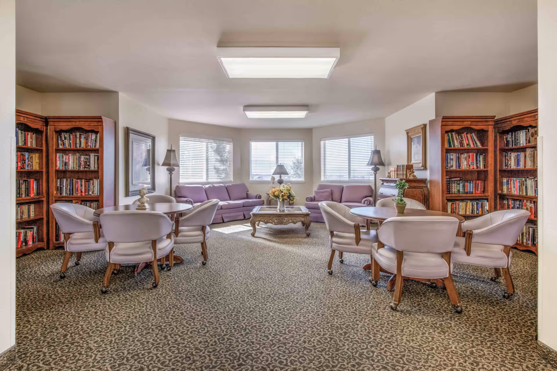 Bright communal lounge with sofas, round tables and chairs, a coffee table, and bookshelves.