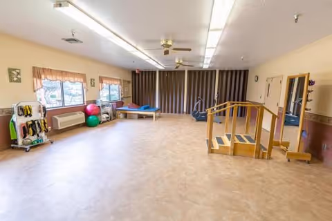 Spacious rehabilitation/activity room with parallel bars, exercise balls, a therapy table, and equipment along the walls.