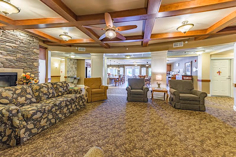A spacious senior living common area with patterned carpet, a floral sofa, three armchairs, a stone fireplace with flowers on the mantel, wooden ceiling beams with ceiling fans, and a view into a dining area with tables and chairs in the background.