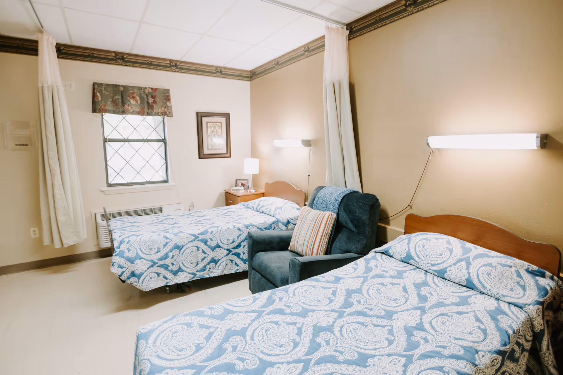 A shared senior living bedroom with two twin beds covered in blue patterned bedspreads, an upholstered chair, and a bedside table under wall-mounted lights.