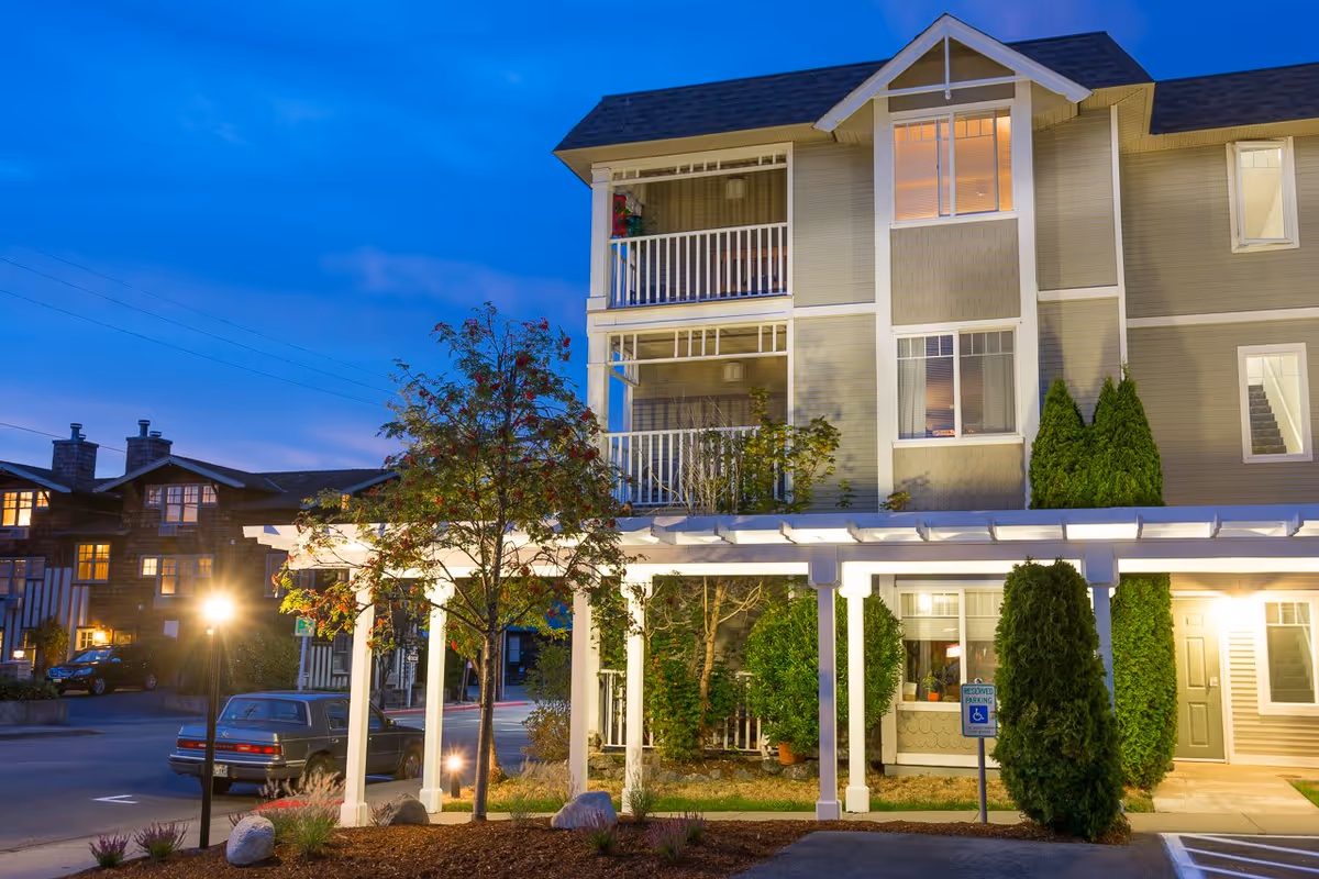 Exterior front of a multi-story senior living building at dusk with lit balconies, a covered walkway, landscaping and parked cars.