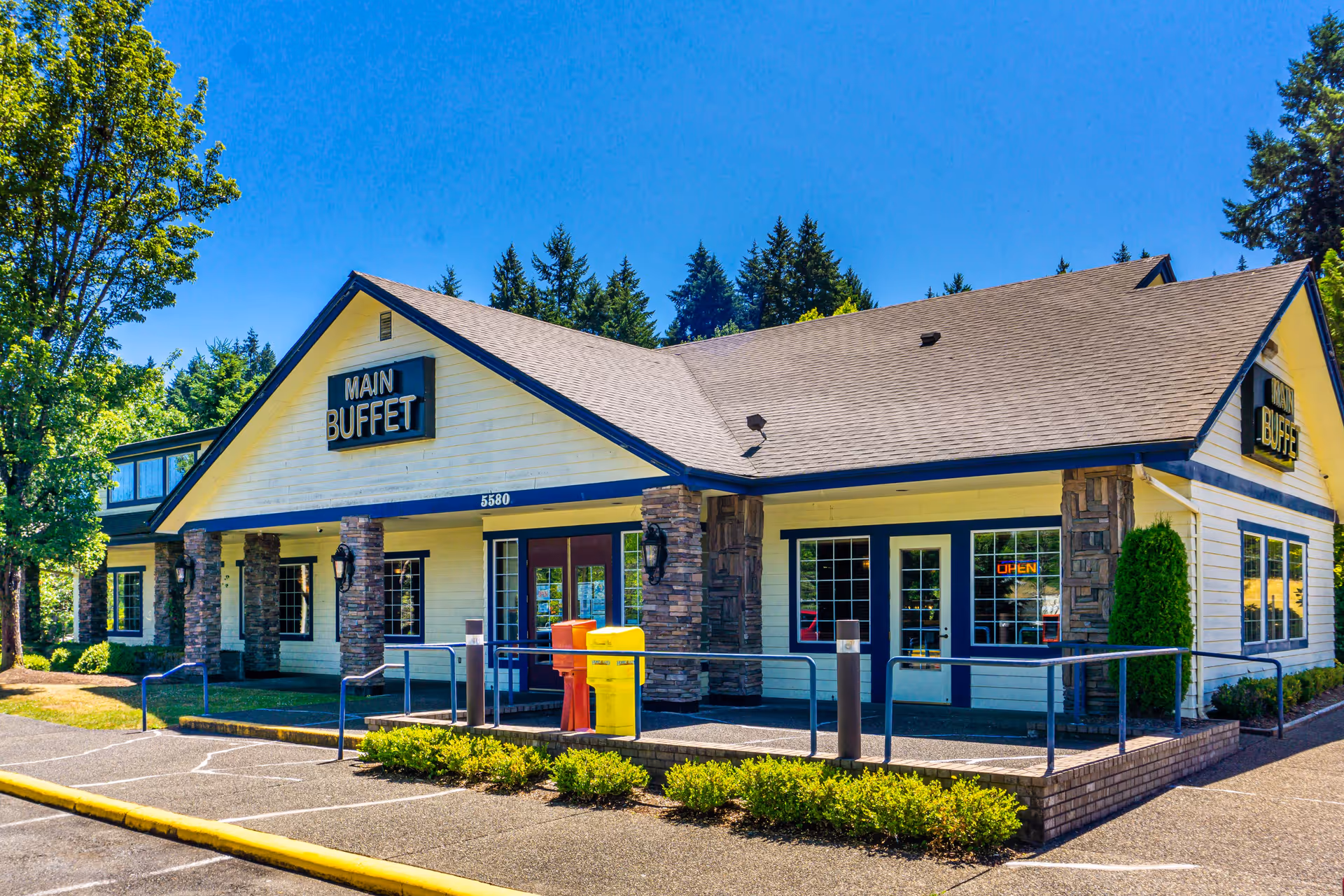 Exterior view of a single-story building with a sign that reads 'MAIN BUFFET'. The building has a beige facade with stone pillars, large windows, and a sloped roof. There are trees and shrubs around the building, and a clear blue sky overhead.