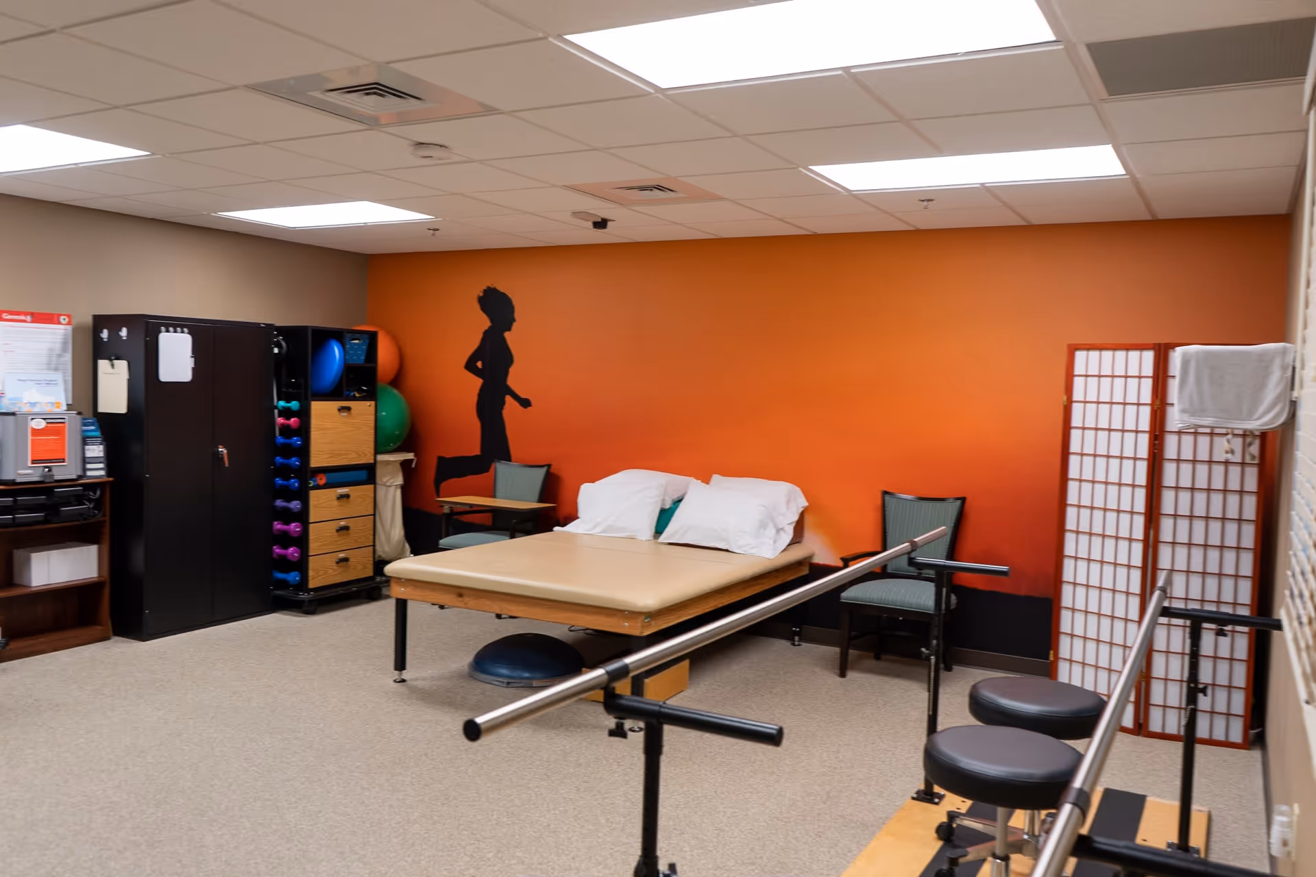 A therapy or rehabilitation room with parallel bars for walking exercises, a padded therapy table with pillows, chairs, storage cabinets with exercise balls and weights, and an orange wall featuring a silhouette of a running person.