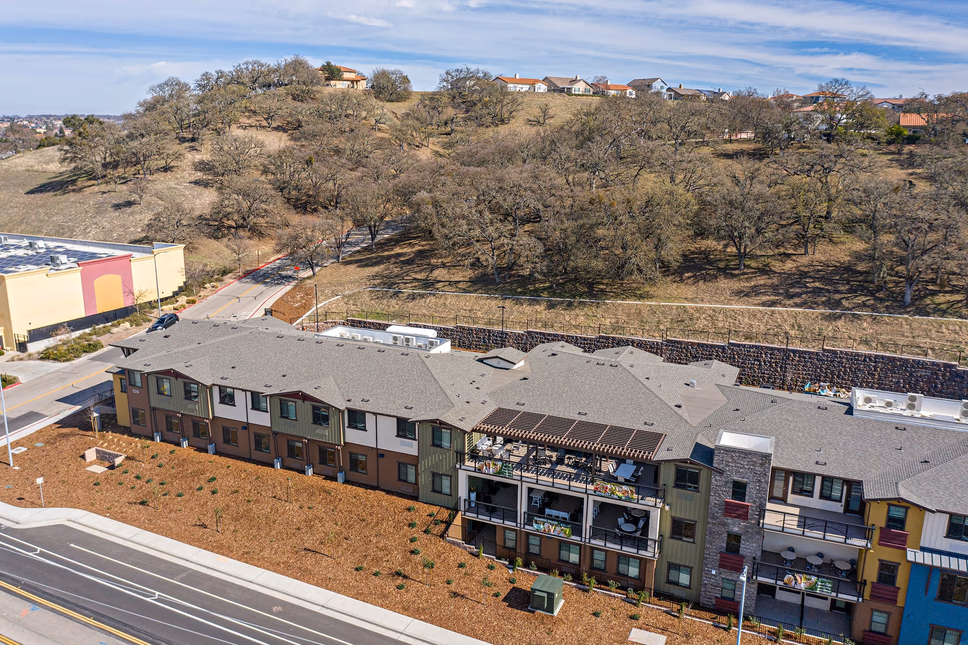 Aerial view of a multi-story senior living facility building with balconies and a rooftop patio area, situated next to a hillside with sparse trees and residential houses in the background under a partly cloudy sky.