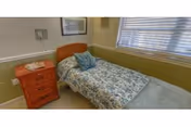Small single-occupancy bedroom with a twin bed covered in a floral bedspread, a wooden nightstand, framed art, and a window with blinds.