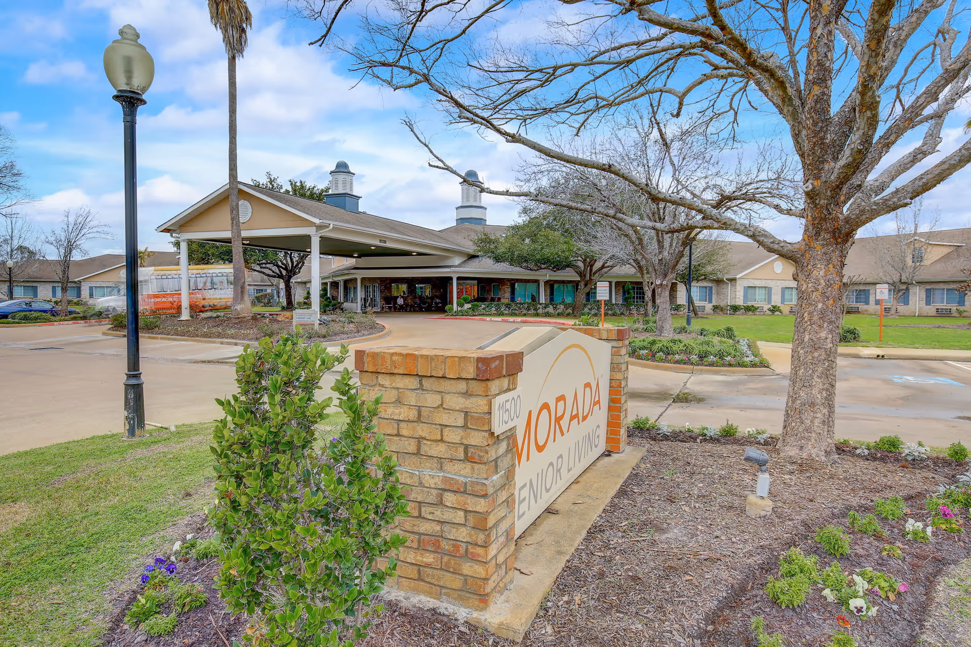 Exterior view of Morada Senior Living facility showing the main entrance with a covered drop-off area, a brick sign with the facility name, landscaped garden beds with flowers and shrubs, a large tree, and a clear blue sky with some clouds.