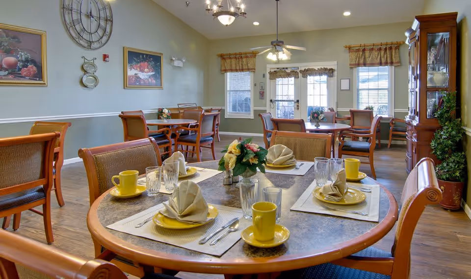 A dining room in a senior living facility with round tables set for four people each. The tables have yellow plates, matching yellow cups, folded beige napkins, silverware, and clear drinking glasses. There are wooden chairs with cushioned seats around the tables. The room has light green walls with framed paintings and a large decorative clock. Windows with valances and a glass door allow natural light to enter. A wooden cabinet and a potted plant are visible in the corner.
