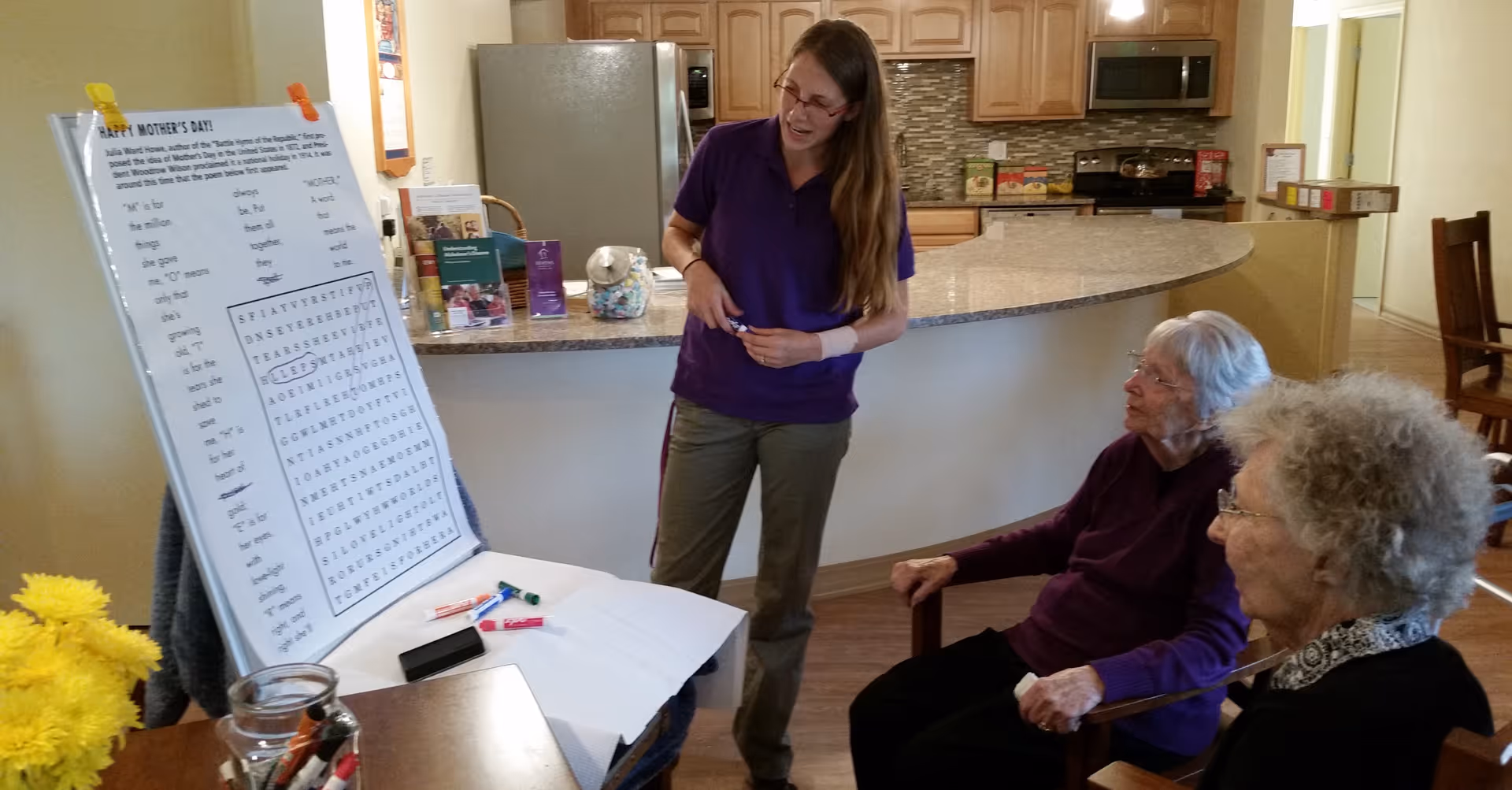 A woman standing and smiling while engaging with two elderly women seated in chairs in a kitchen area. The standing woman is pointing to a large word search puzzle on an easel that has a Mother's Day theme. The kitchen has wooden cabinets, a refrigerator, and a microwave. There are yellow flowers and markers on a nearby table.