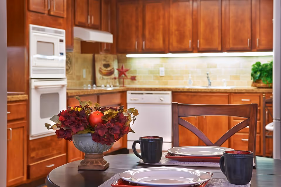 A kitchen with wooden cabinets, a built-in oven and microwave, a dishwasher, and a countertop with a sink. In the foreground, there is a round dining table set with two place settings including plates and black mugs, and a decorative flower arrangement with red and yellow flowers and a red apple.