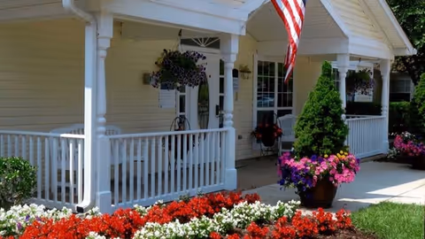 Front porch of a light-colored building with white columns and railing, hanging baskets and potted flowers, an American flag, and a colorful flower bed.