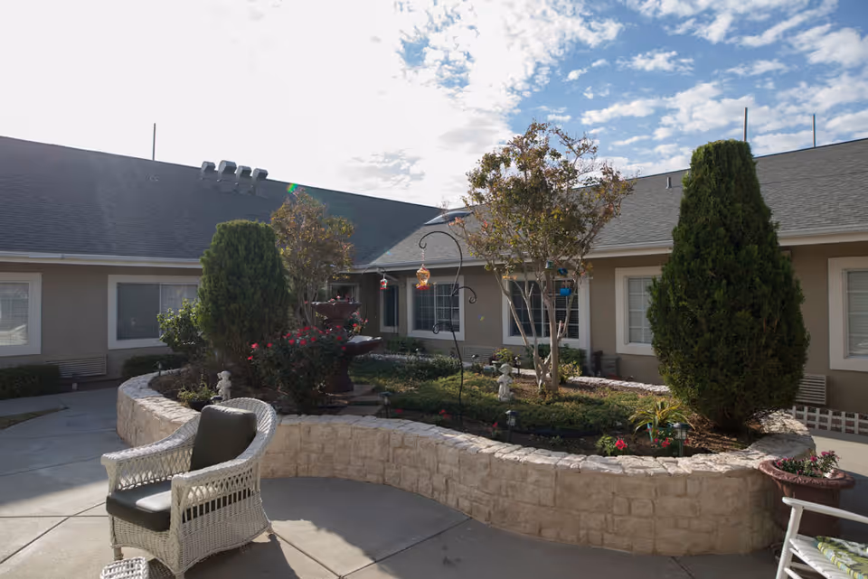 Outdoor courtyard area at SunRidge at Desert Springs featuring a stone raised garden bed with various plants and small trees, a decorative fountain, bird feeders, and white wicker chairs with cushions on a concrete patio under a partly cloudy sky.