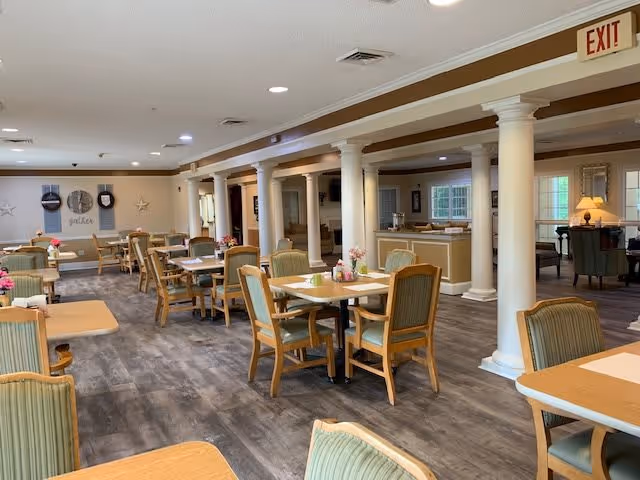 A spacious dining room in a senior living facility with multiple wooden tables and green cushioned chairs arranged neatly. The room features white columns, wood flooring, and soft lighting. Decorative elements include wall art and small flower arrangements on the tables.