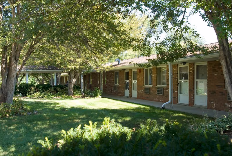 Exterior view of a single-story brick building with several doors and windows, surrounded by green grass, bushes, and large trees providing shade.