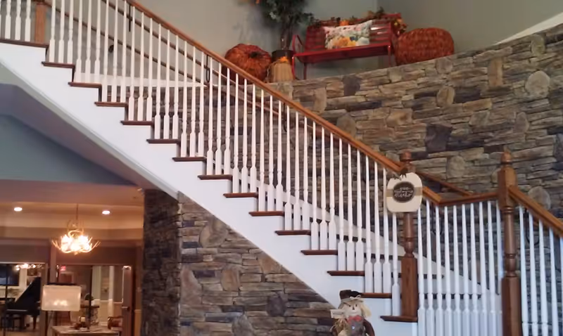 Interior view of a senior living facility showing a staircase with white spindles and wooden handrails against a stone wall. There are decorative items on the ledge above the stone wall, including baskets and a red bench with pillows. A scarecrow decoration is placed at the base of the stairs. In the background, there is a chandelier and part of a room with furniture visible.