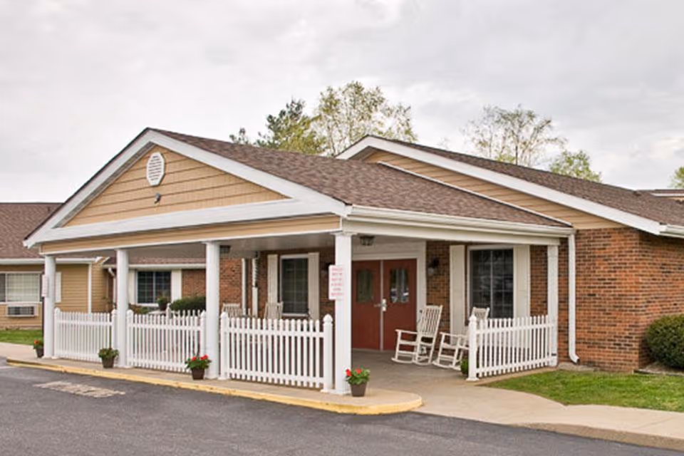 Exterior view of Mitchell Manor, showing a single-story building with a covered entrance supported by white columns, white picket fencing, potted plants, and two rocking chairs on the porch.