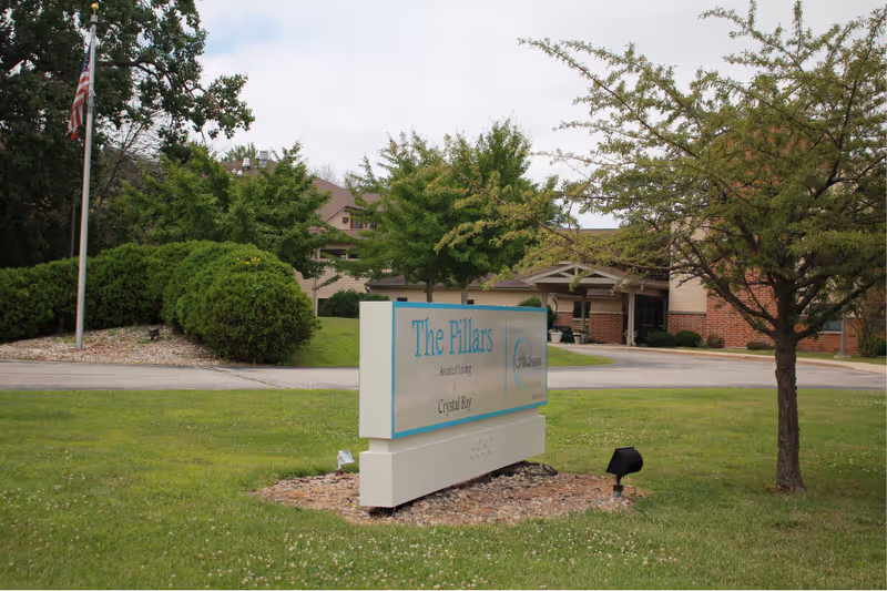 A freestanding sign reading "The Pillars" on a grassy lawn in front of the entrance to a senior living facility with trees and a driveway.