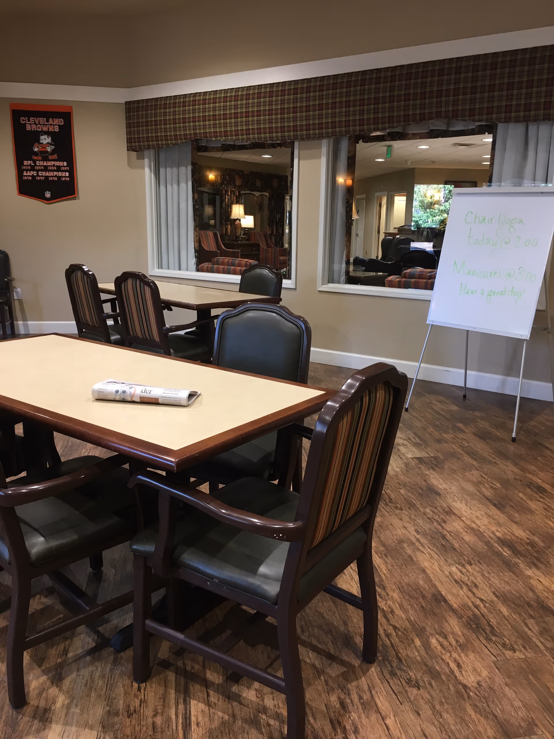 Communal dining/activity room with tables and chairs, a newspaper on a table, a whiteboard easel, and windows into a lounge.