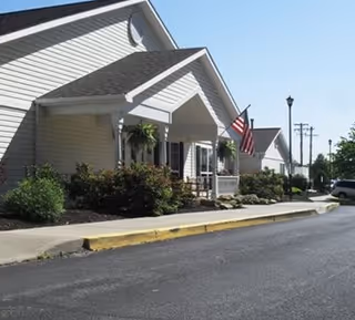 Exterior view of a single-story residential building with beige siding, a covered porch with hanging plants, an American flag, and a paved driveway with a yellow curb.