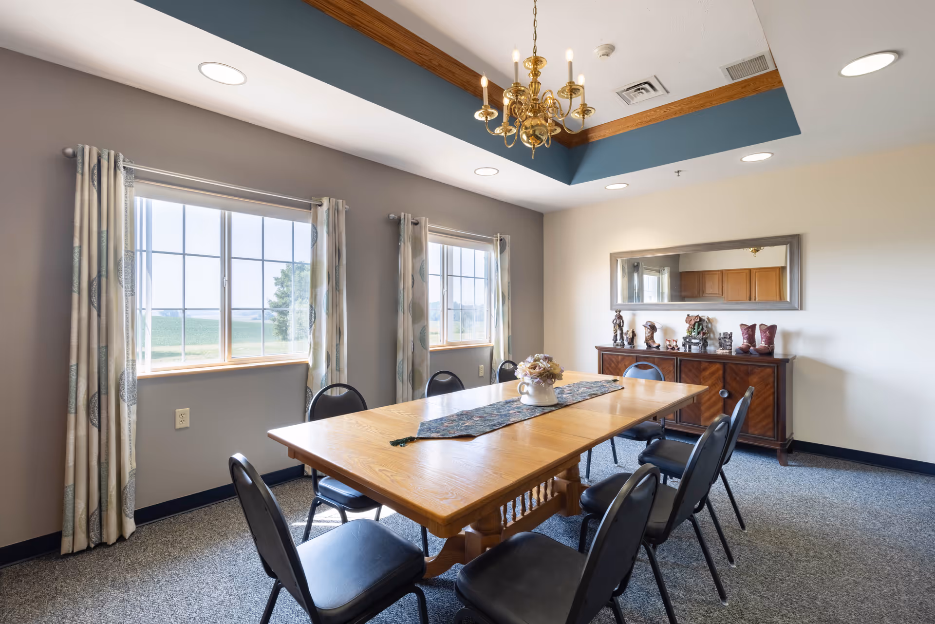 A dining room with a large wooden table surrounded by eight black chairs. The room has two large windows with patterned curtains, a decorative chandelier hanging from a tray ceiling with wood trim, and a sideboard with various decorative items and a large mirror above it.