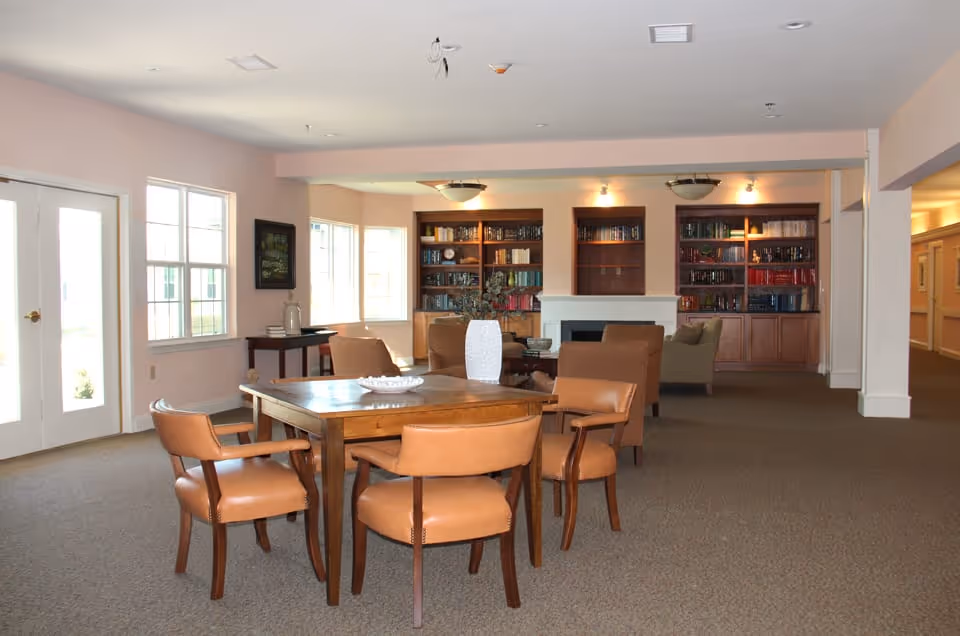 A spacious common area in a senior living facility featuring a wooden table with four brown leather chairs in the foreground. In the background, there are several armchairs arranged around a fireplace with built-in bookshelves filled with books on either side. Large windows allow natural light to brighten the room, and the walls are painted a soft beige color.