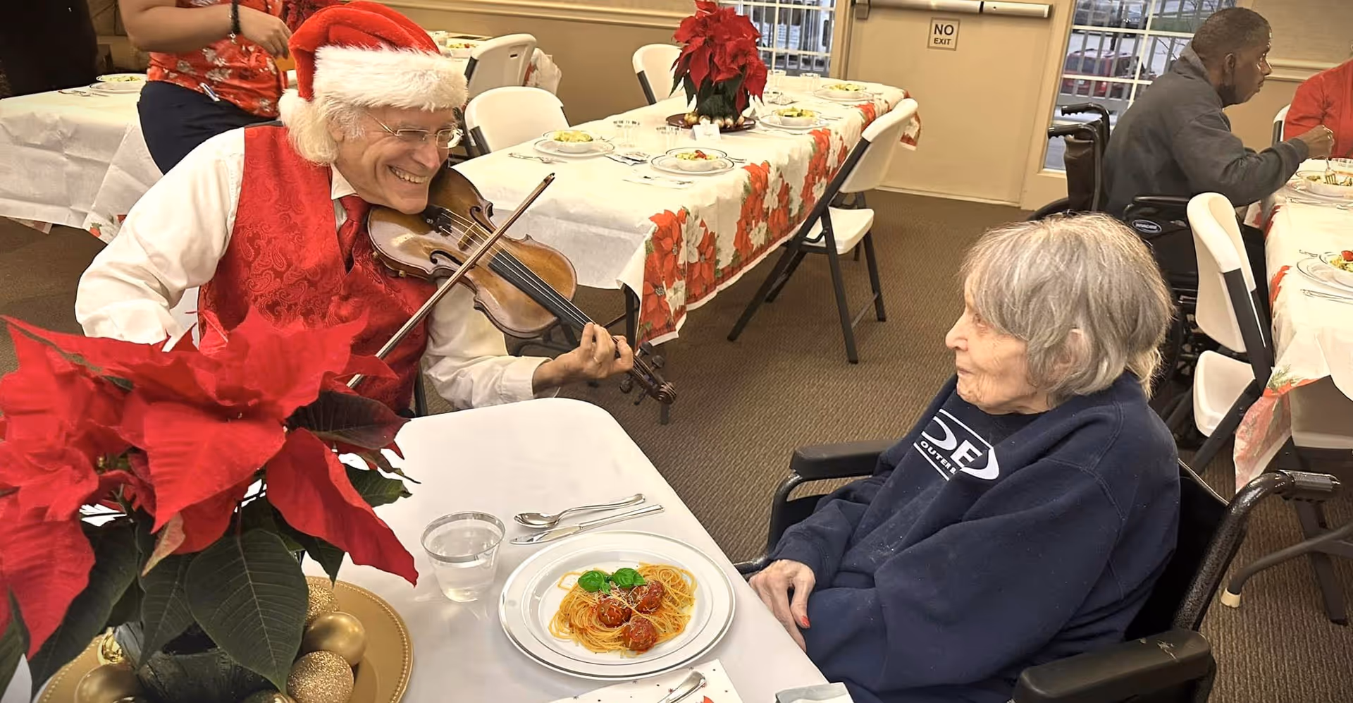 A man in a Santa hat plays the violin for a seated elderly woman at a holiday-themed dining table.