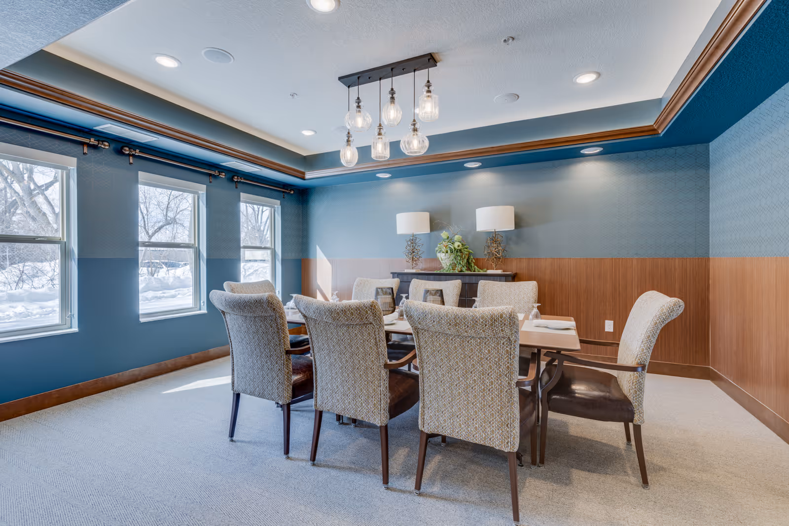 A dining room with a rectangular table surrounded by eight upholstered chairs. The walls are painted blue with wooden paneling on the lower half. There are three windows on the left side letting in natural light, and a modern chandelier with multiple hanging light bulbs above the table. Two table lamps and a plant are on a sideboard against the far wall.