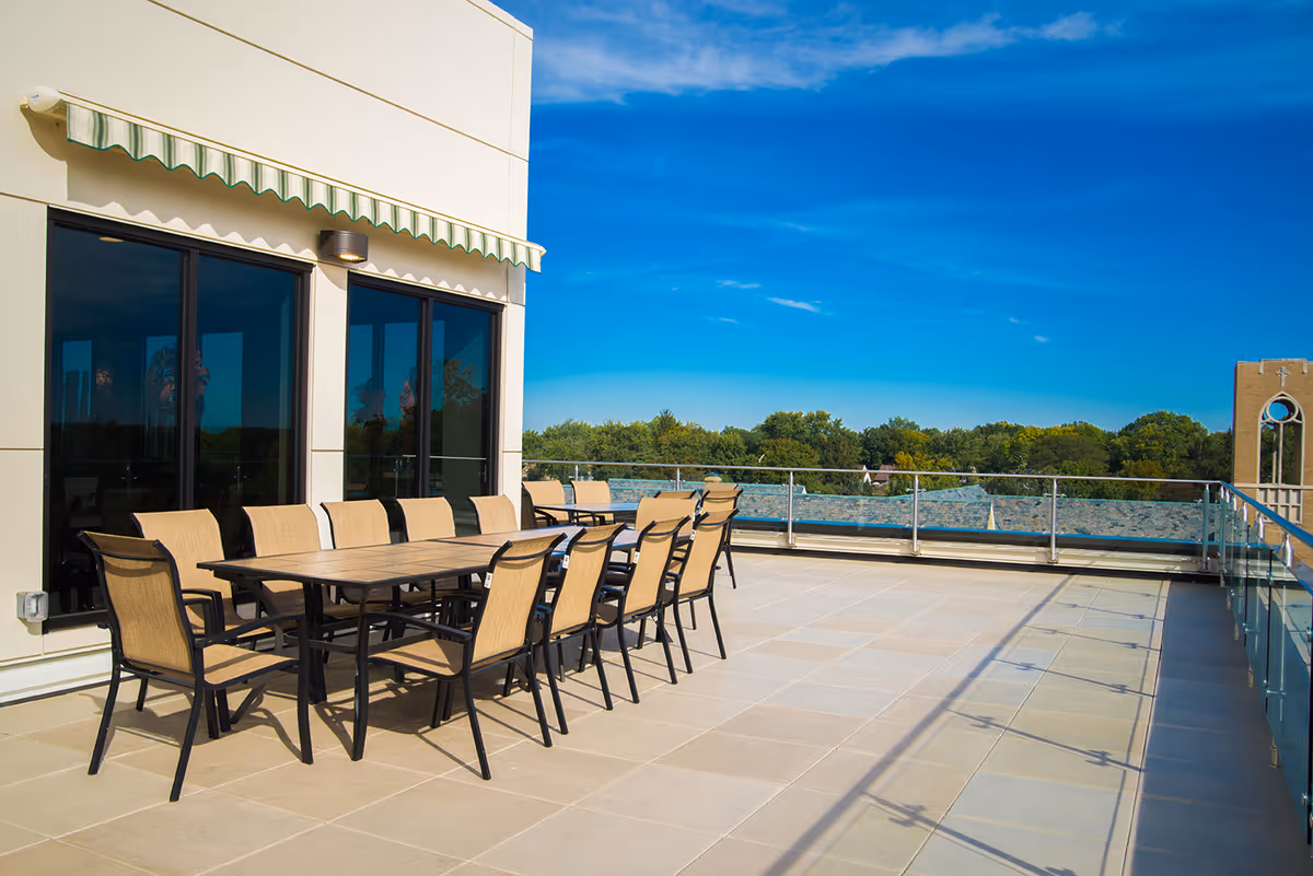 Outdoor terrace with a long rectangular table surrounded by twelve beige and black chairs. The terrace has a glass railing and overlooks a green tree line under a clear blue sky. The building wall has large windows and a green and white striped awning.