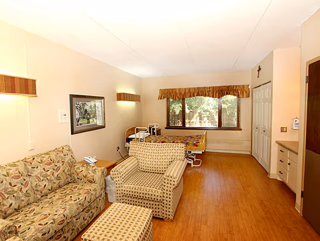 Bright studio-style senior living bedroom with a hospital-style bed, sofa, armchair with ottoman, and a large window.