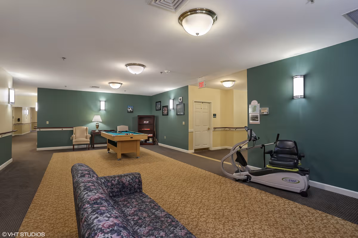 Interior common area with green and beige walls, featuring a floral patterned couch, a pool table, a beige armchair with a side table and lamp, a bookshelf, and a NuStep exercise machine. The area is well-lit with ceiling and wall lights, and there is an exit door in the background.