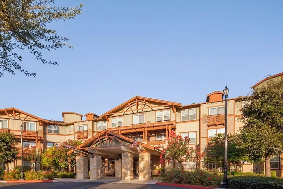 Exterior view of a multi-story senior living facility building with a covered entrance, surrounded by trees and landscaping under a clear blue sky.
