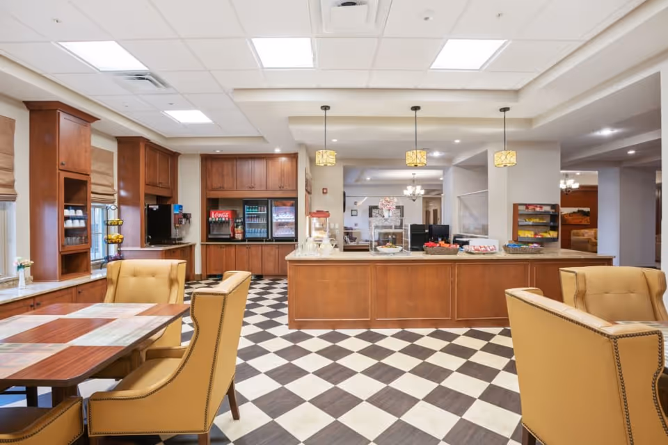A bright and spacious dining area with checkered black and white tile flooring, wooden cabinetry, and a long counter with snacks and beverages. There are comfortable yellow upholstered chairs around wooden tables, pendant lights hanging from the ceiling, and windows with beige Roman shades.