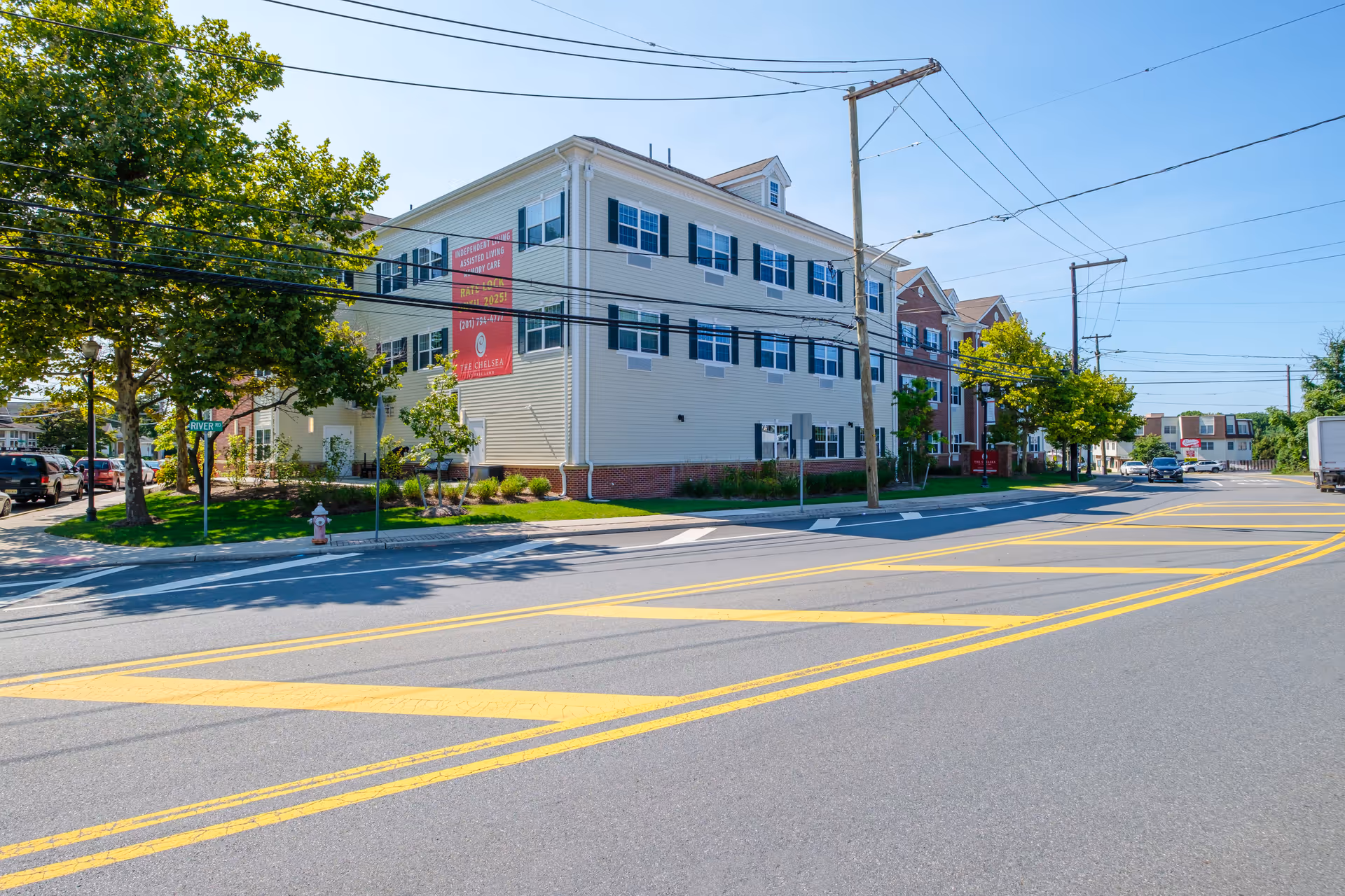 Exterior view of The Chelsea at Fair Lawn, a multi-story senior living facility with beige and red brick siding, multiple windows with black shutters, and a large red banner advertising independent living, assisted living, and memory care. The building is situated along a street with yellow road markings, trees, and parked cars.