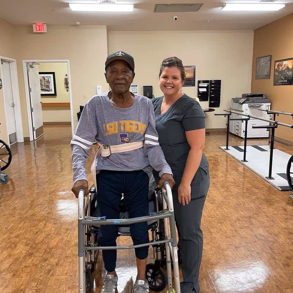 An elderly man using a walker is standing next to a smiling female caregiver in a healthcare facility hallway. The man is wearing a LSU Tigers shirt and a cap, and the caregiver is dressed in gray scrubs. The hallway has wooden flooring, handrails, a printer, and motivational posters on the walls.