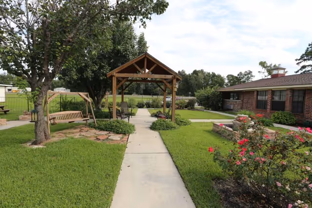 Outdoor garden area with a concrete walkway leading to a wooden gazebo. There is a wooden swing on the left side under a tree and a flower bed with blooming pink flowers on the right. A brick building is visible on the right side of the image.