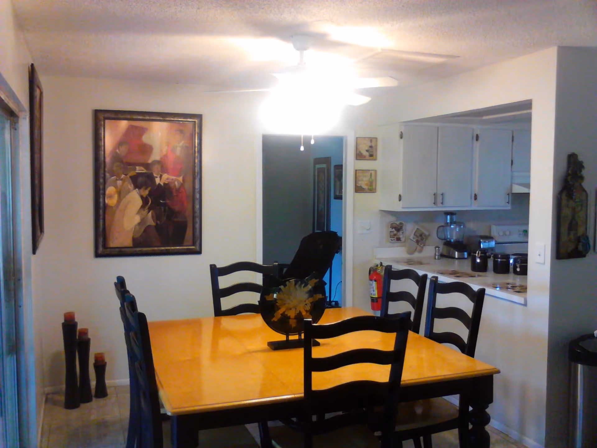 Dining room with a wooden table and six chairs under a ceiling fan light, wall art on the left wall, and a pass-through opening to a white kitchen.