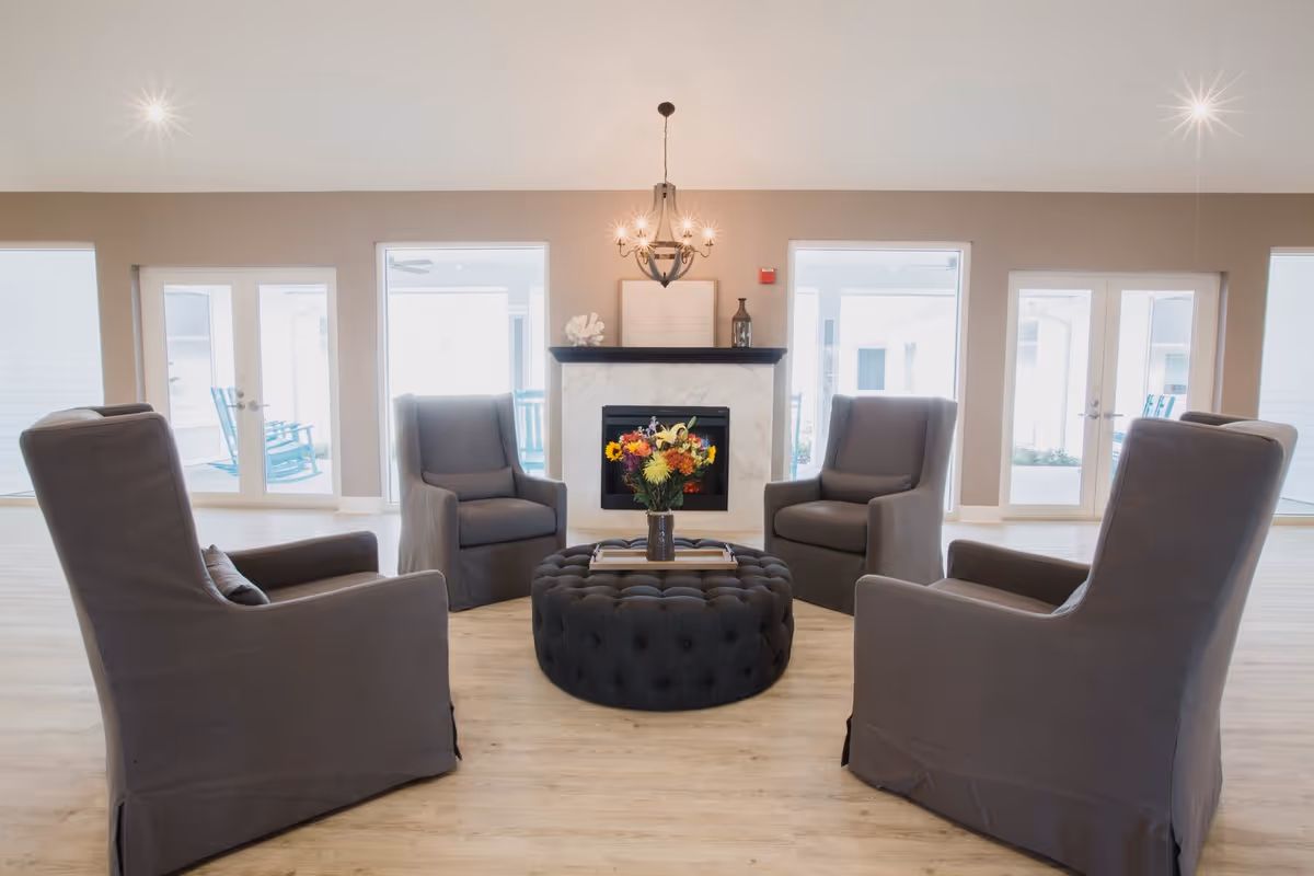 A bright and spacious living room area with four gray upholstered armchairs arranged in a circle around a large, round, tufted ottoman with a tray and a vase of colorful flowers on top. The room features a fireplace with a marble surround and a decorative chandelier hanging from the ceiling. Large windows and glass doors allow natural light to fill the space, with views of outdoor rocking chairs.