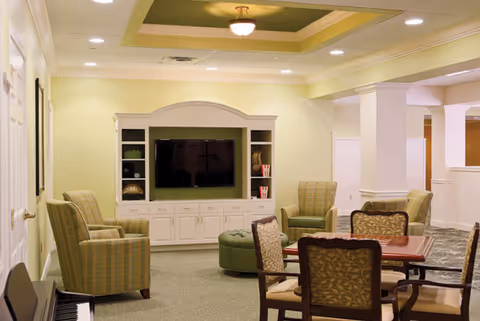 A cozy living room area in a senior living facility featuring a built-in entertainment center with a TV, surrounded by shelves with decorative items. The room has several upholstered armchairs and a round ottoman, along with a table and chairs in the foreground. The ceiling has a recessed design with a central light fixture, and the walls are painted a soft yellow color.