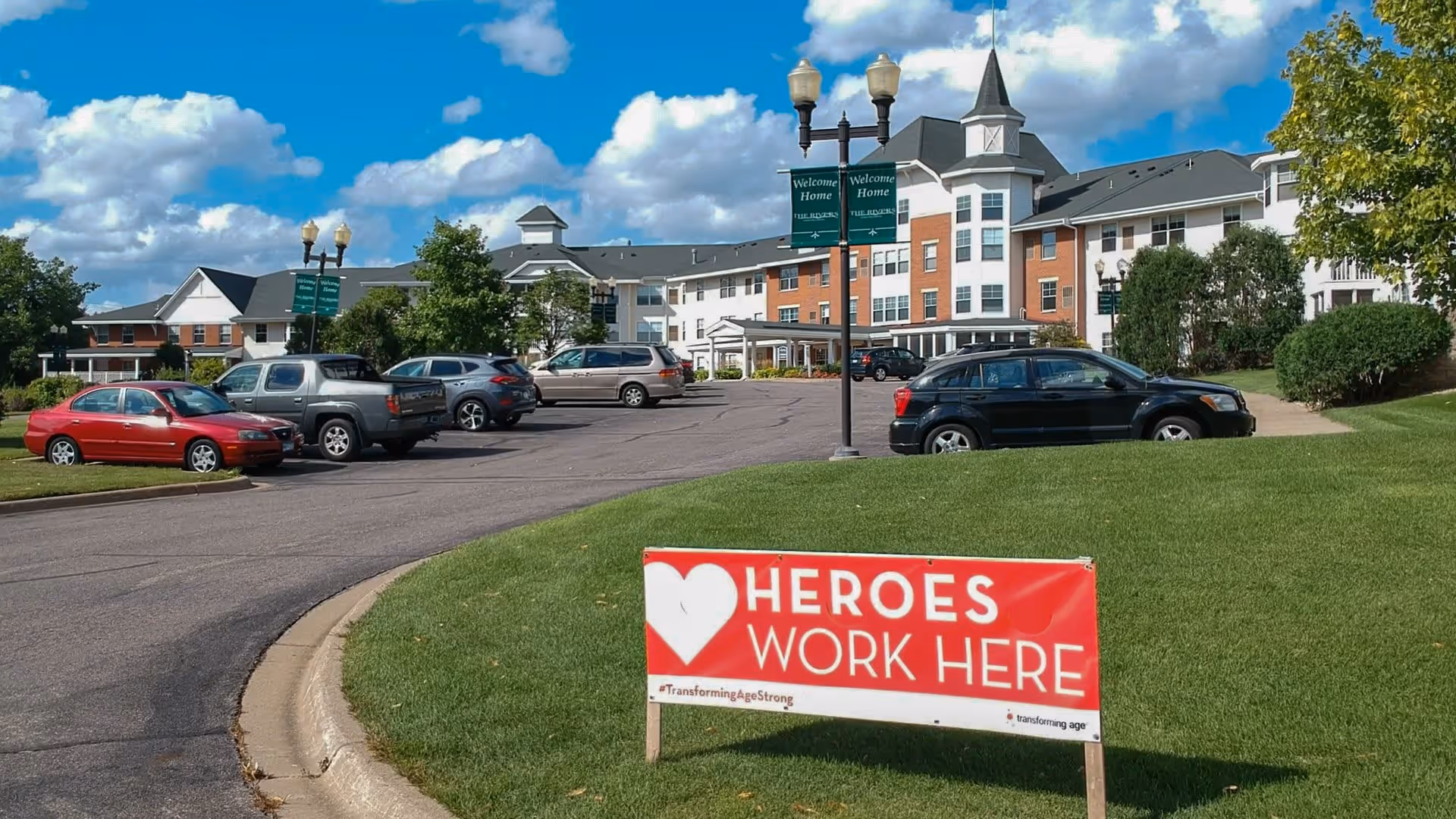 Exterior view of The Rivers Retirement Community building with a parking lot in front. Several cars are parked along the driveway, and a red and white sign on the grass reads 'HEROES WORK HERE' with a heart symbol. The sky is partly cloudy with blue patches visible.