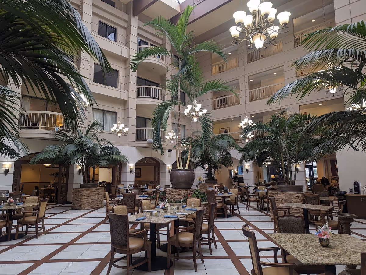 Spacious indoor dining area with multiple tables and chairs arranged neatly. Large potted palm trees are placed throughout the space, and elegant chandeliers hang from the high ceiling. The area is surrounded by multiple floors with balconies overlooking the dining space.