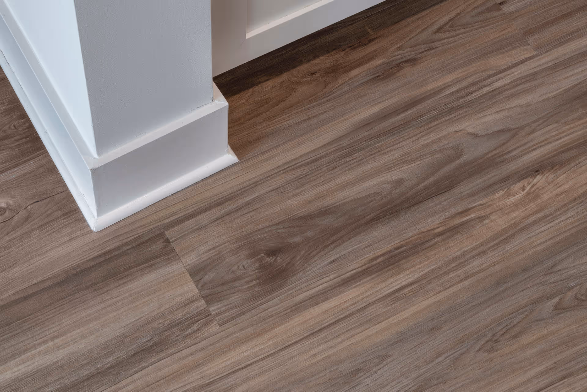 Close-up view of a corner of a white wall with baseboard trim and wood-patterned flooring in shades of brown and gray.