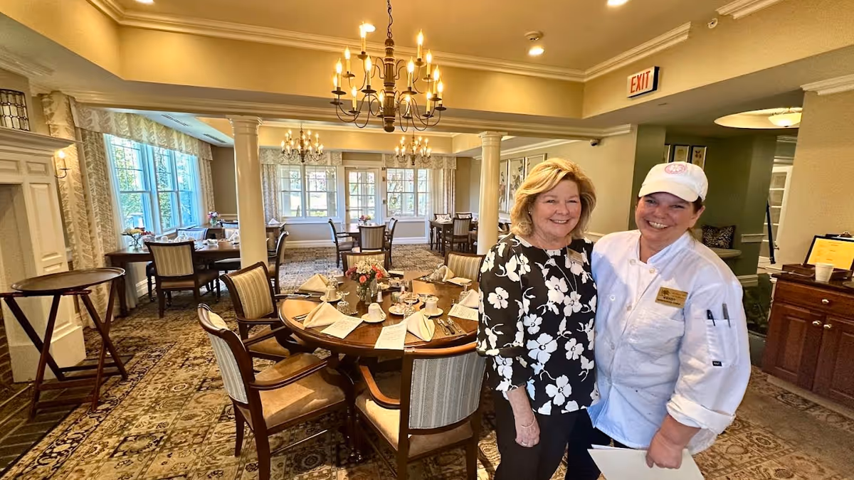 Two smiling women standing in a warmly lit dining room with round tables set with napkins, cups, and flowers. The room features large windows with curtains, chandeliers, and patterned carpet. One woman is wearing a floral blouse and the other is dressed in a white chef's uniform and cap.